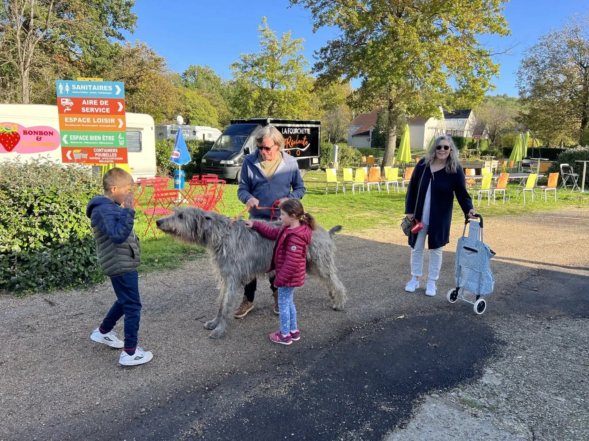 Eine Familie interagiert mit einem großen Hund in der Nähe eines Schildes mit verschiedenen Wegweisern und Dienstleistungen.