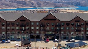 Un hotel de varios pisos con techo plano, ventanas grandes y vista a un lago y montañas al fondo.