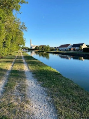 A calm river with a gravel path, houses, and a tall chimney in the background. - ZEcamping 3 étoiles dog friendly locatif 2 pers (Bagneaux-sur-Loing)