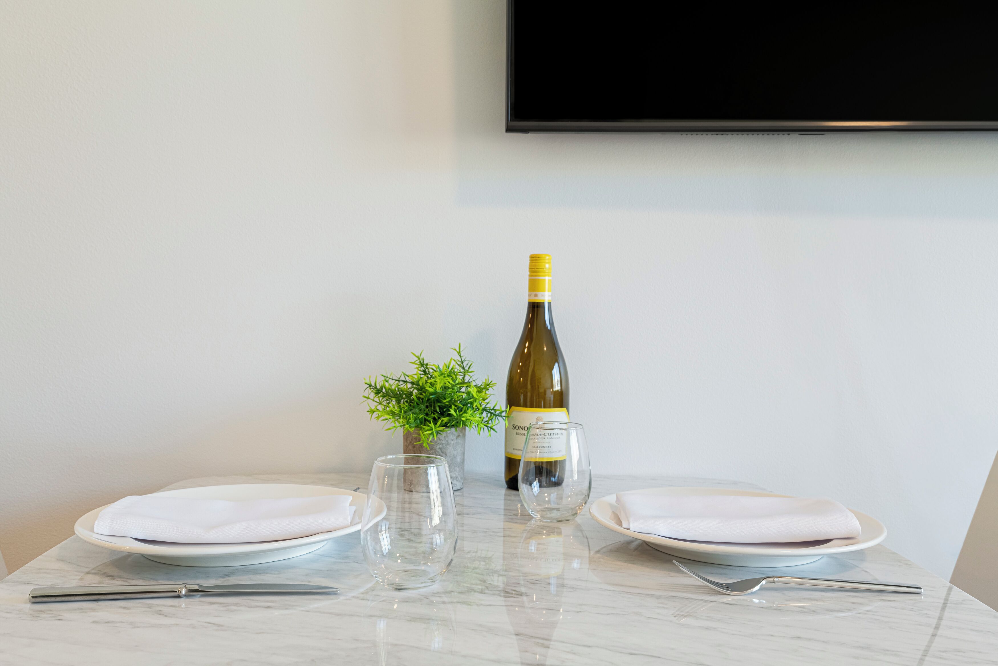 Una mesa con dos platos blancos, dos copas de vino vacías, una botella de vino y una pequeña planta en maceta.