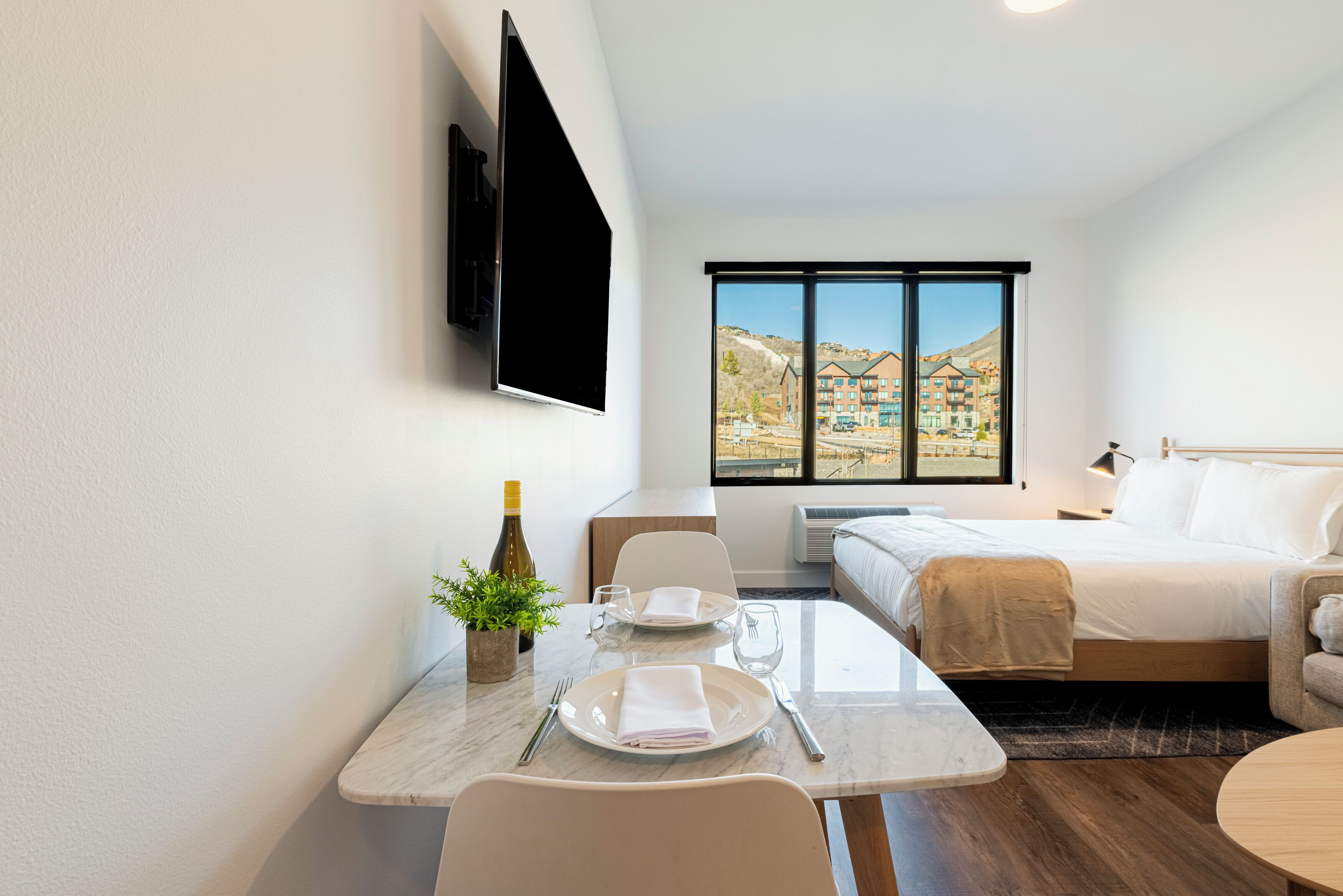 Una camera d'albergo con un letto, una televisione fissata al muro, un tavolo da pranzo con sedie e una finestra che si affaccia su edifici.