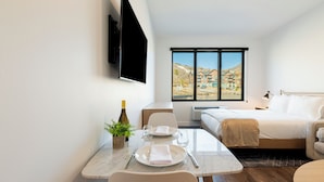 Una camera d'albergo con un letto, una televisione fissata al muro, un tavolo da pranzo con sedie e una finestra che si affaccia su edifici.