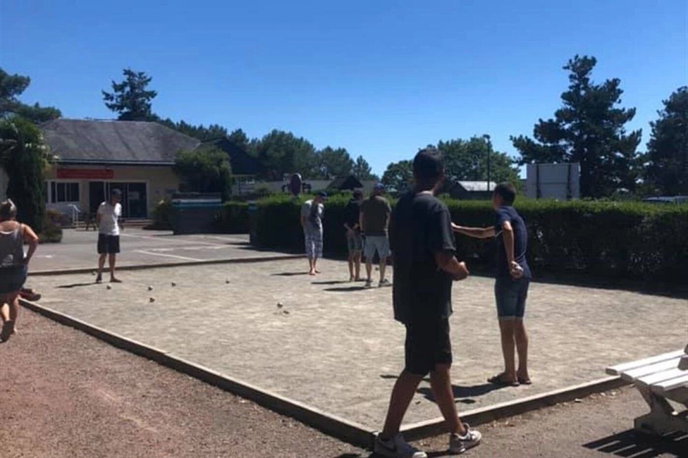 Een groep mensen speelt jeu de boules in een open gebied met een verhard oppervlak, omringd door bomen en een gebouw op de achtergrond.