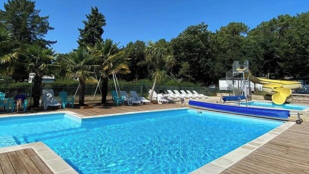 A swimming pool with a wooden deck, lounge chairs, and palm trees in the background. - ZEcamping 3* animé piscine chauffée animaux acceptés locatif 17m2 (Guémené-Penfao)