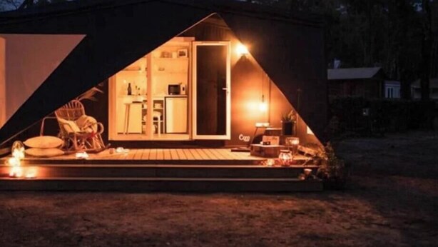 Une petite cabane de forme triangulaire, avec un espace cuisine visible, une table à manger et une chaise à l’extérieur.