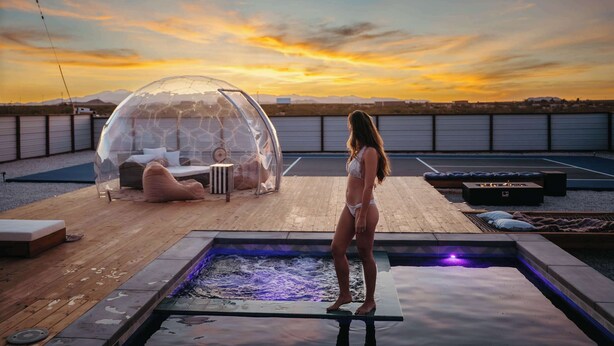 Una mujer de pie junto a una piscina con una estructura en forma de cúpula transparente y una zona de estar en una terraza en la azotea.