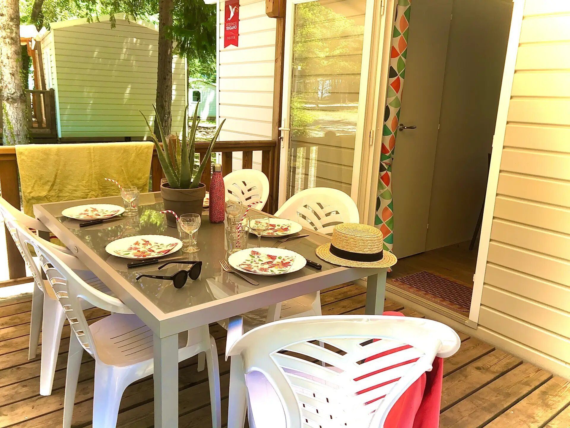 Uma área de refeições ao ar livre com uma mesa posta para quatro pessoas, cercada por cadeiras de plástico branco, um vaso de planta e um chapéu decorativo sobre a mesa.