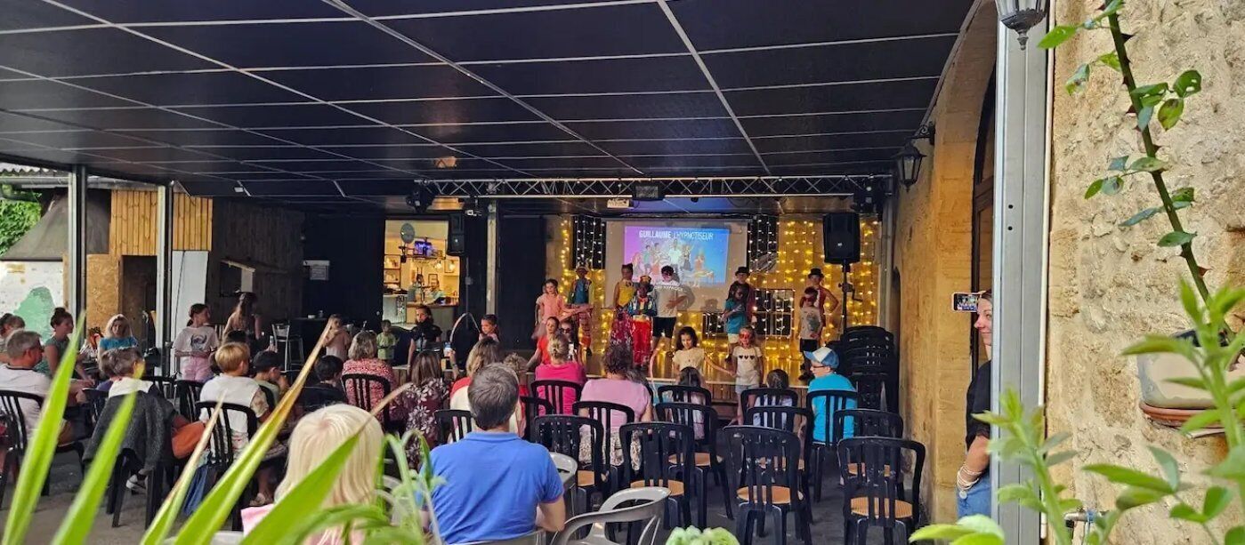 An outdoor seating area with tables and chairs, a stage with performers, and a backdrop with text.
