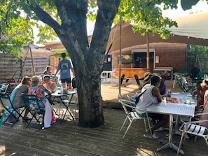 An outdoor dining area with tables and chairs under a large tree, a wooden building in the background, and a covered seating area. - ZEcamping 3 étoiles piscine chauffée tente 18m2 (Lussas)