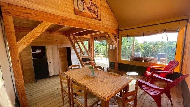 A wooden interior with a dining table, chairs, a bicycle artwork, and a view through the windows. - ZEcamping 3* piscine chiens acceptés locatif BBQ 72m2 5 pers (Dévillac)