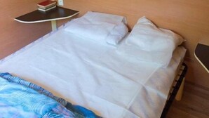 Uma cama de solteiro com roupa de cama branca, uma pequena mesa de cabeceira com um livro, uma janela com cortinas e um chão de madeira.