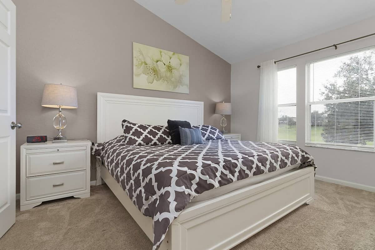 A bedroom with a bed, nightstands, lamps, a wall clock, and a window with blinds.