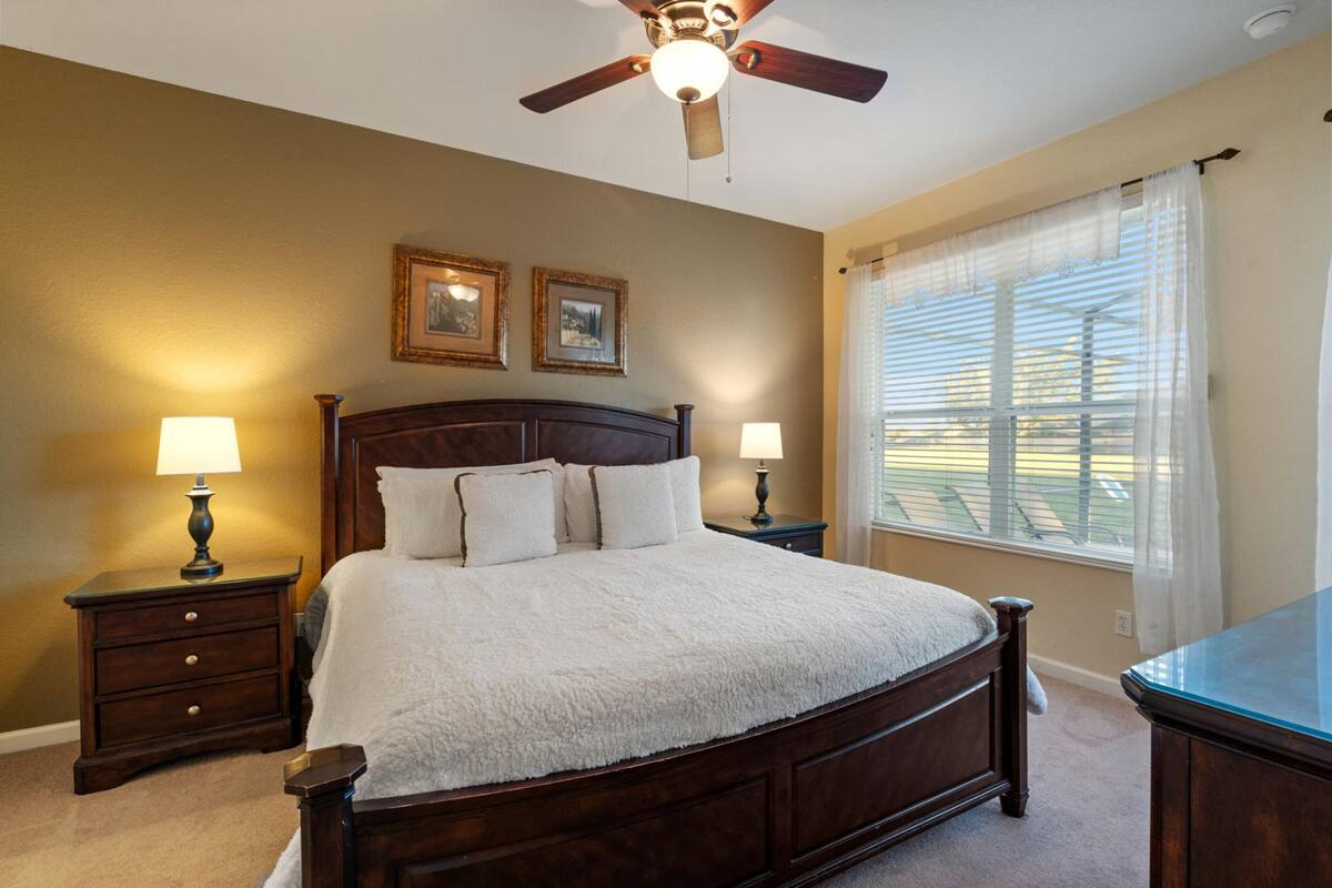 A bedroom with a large bed, two bedside tables with lamps, a ceiling fan, and a window with blinds.