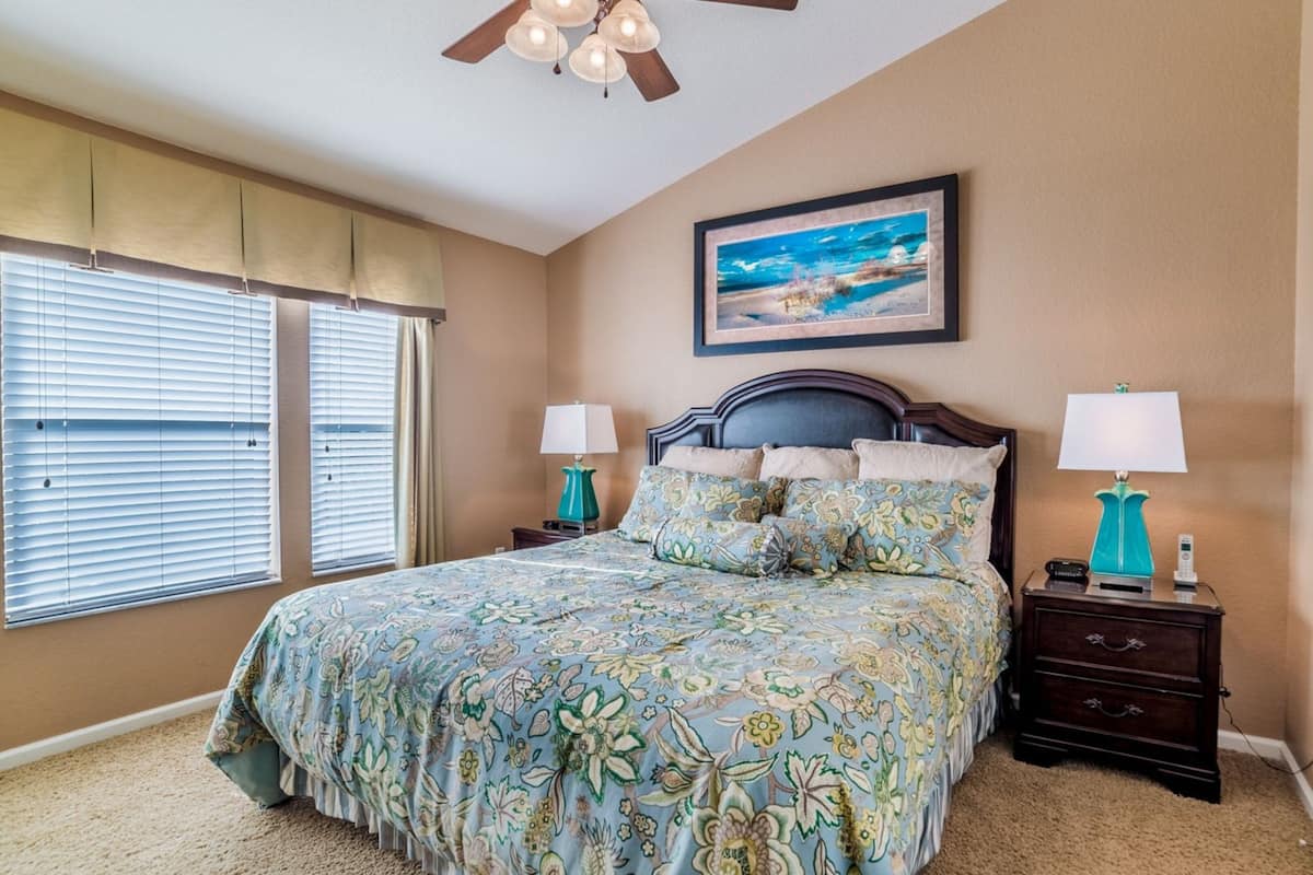 A bedroom with a bed, two bedside tables, a ceiling fan, a window with blinds, and a framed picture on the wall.