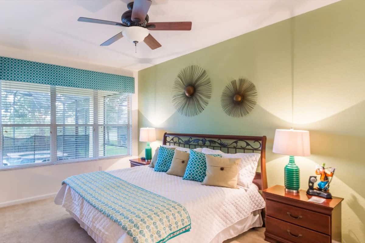 A bedroom with a bed, nightstands, a ceiling fan, a window with blinds, and decorative wall art.