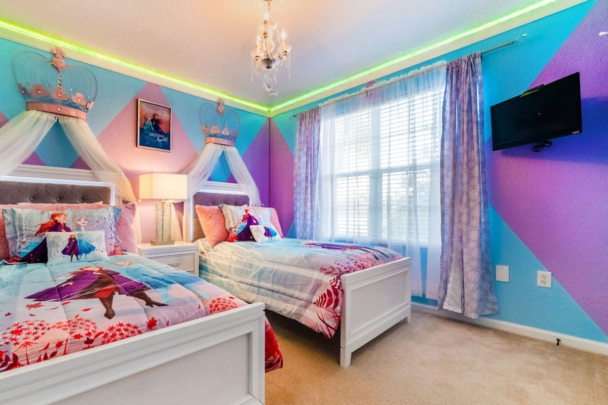 A bedroom with two beds, a chandelier, a TV, and a large window with curtains.