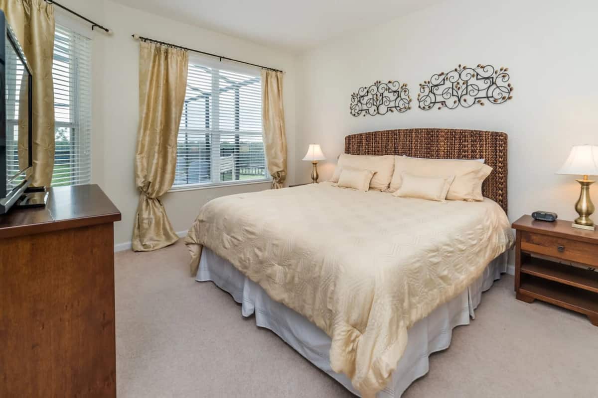 A bedroom with a large bed, two bedside tables with lamps, a TV on a stand, and a window with curtains.