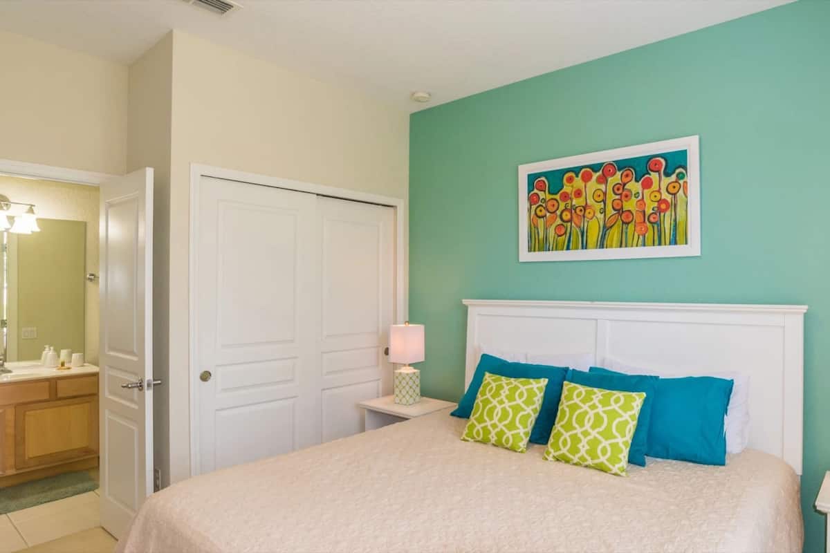 A bedroom with a bed, a nightstand, a bedside lamp, a painting on the wall, and a door to another room.