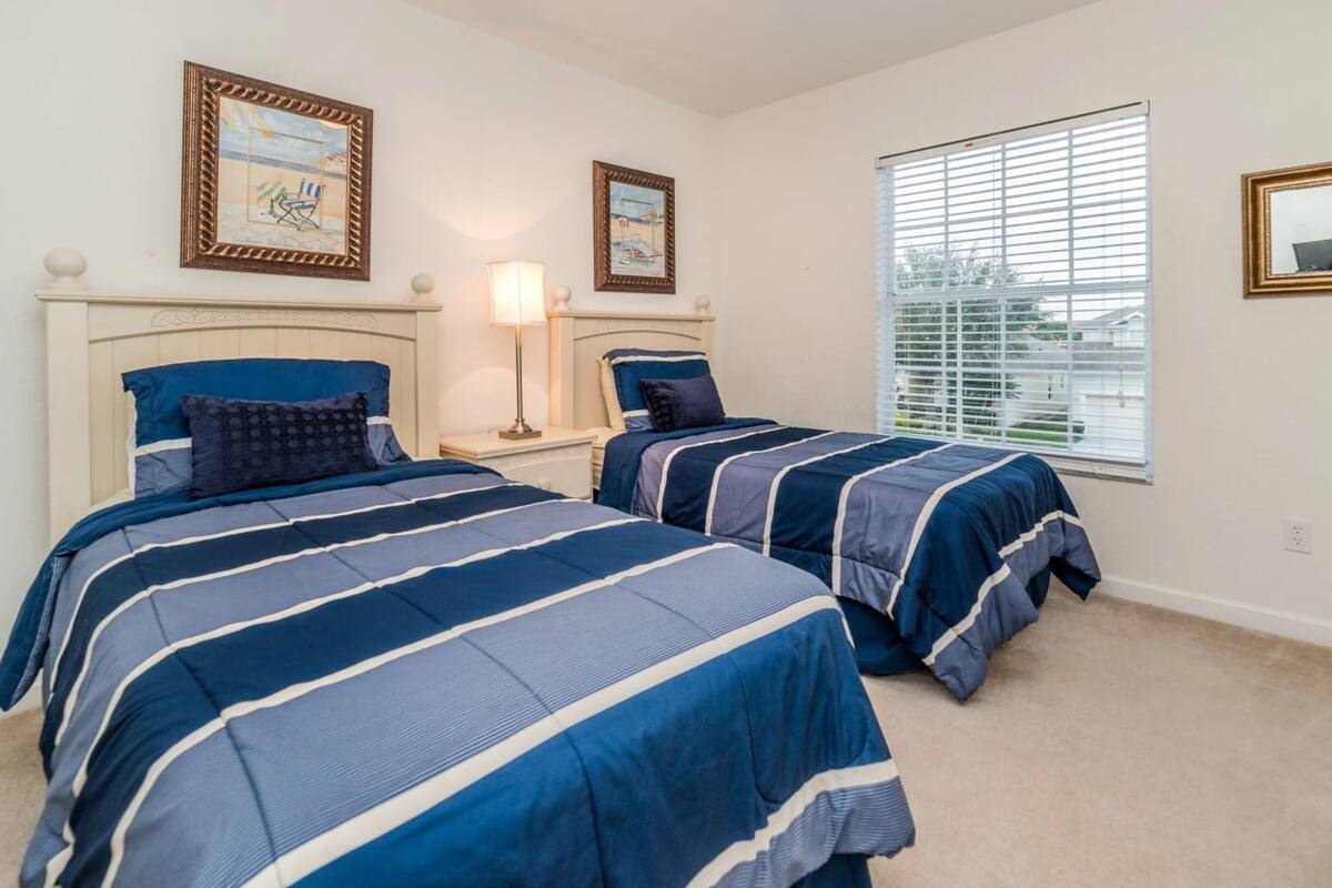 A bedroom with two beds, a nightstand, a lamp, a window with blinds, and framed pictures on the wall.