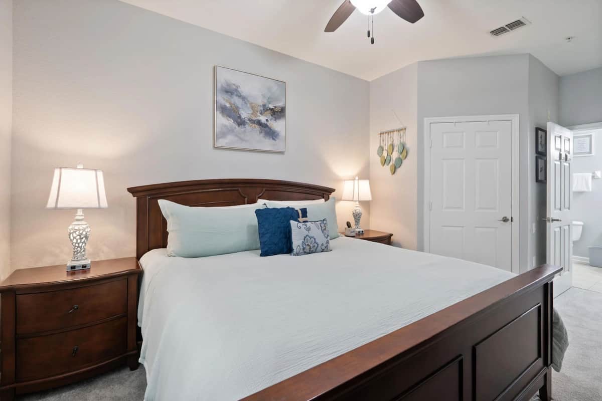 A bedroom with a bed, two bedside tables, a ceiling fan, a door, and a wall-mounted artwork.