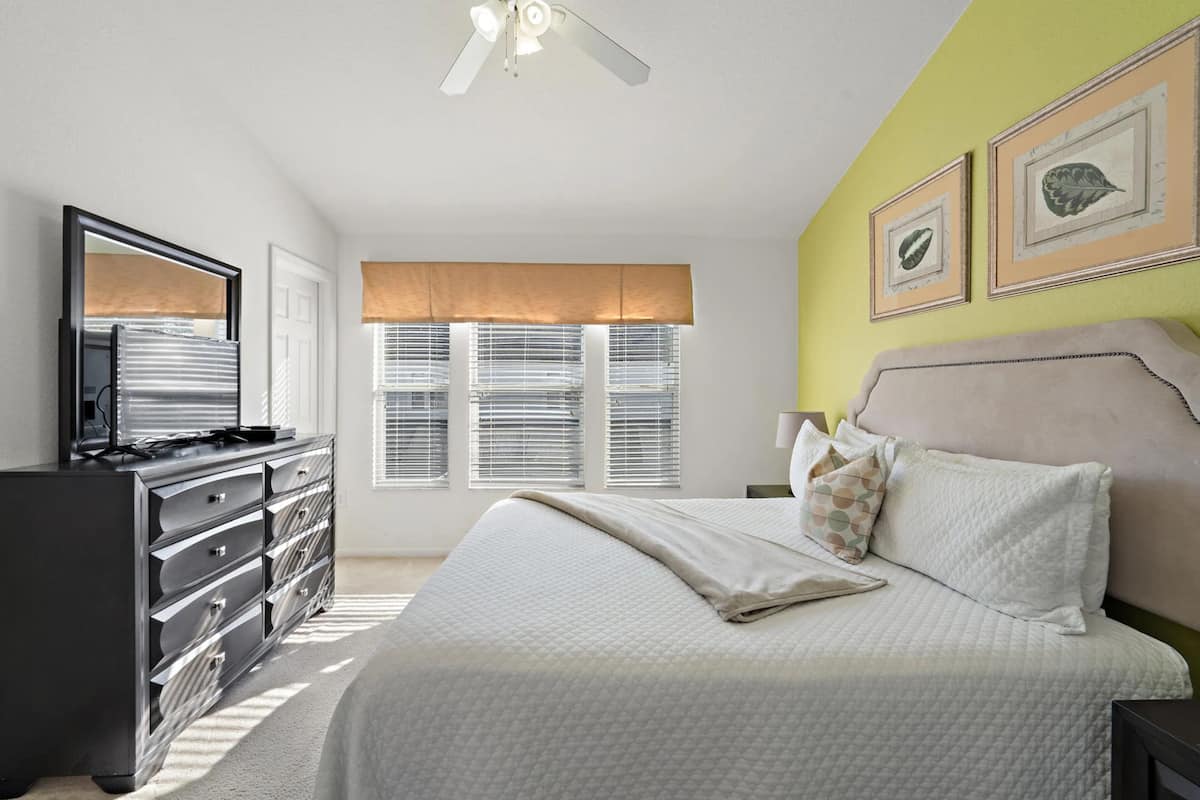 A bedroom with a bed, dresser, ceiling fan, windows with blinds, and wall art.
