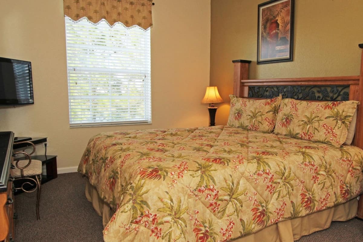 A bedroom with a bed, a TV, a window with blinds, a nightstand, and a lamp.