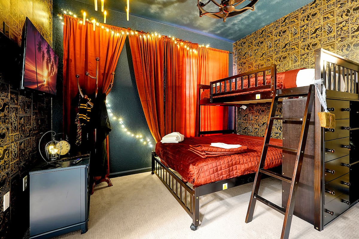 A bunk bed with a ladder, a desk with a clock, and a TV on the wall.