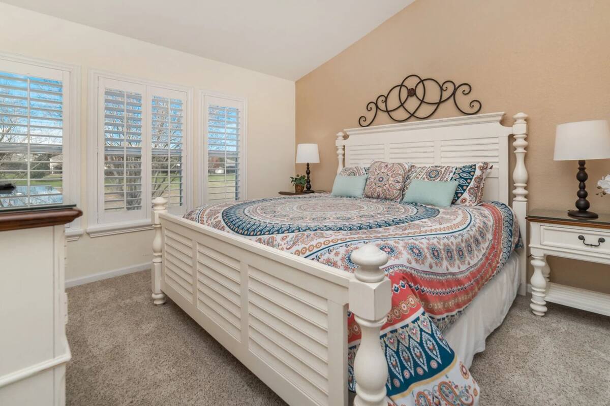 A bedroom with a white bed, two bedside tables, a window with blinds, and a beige wall.