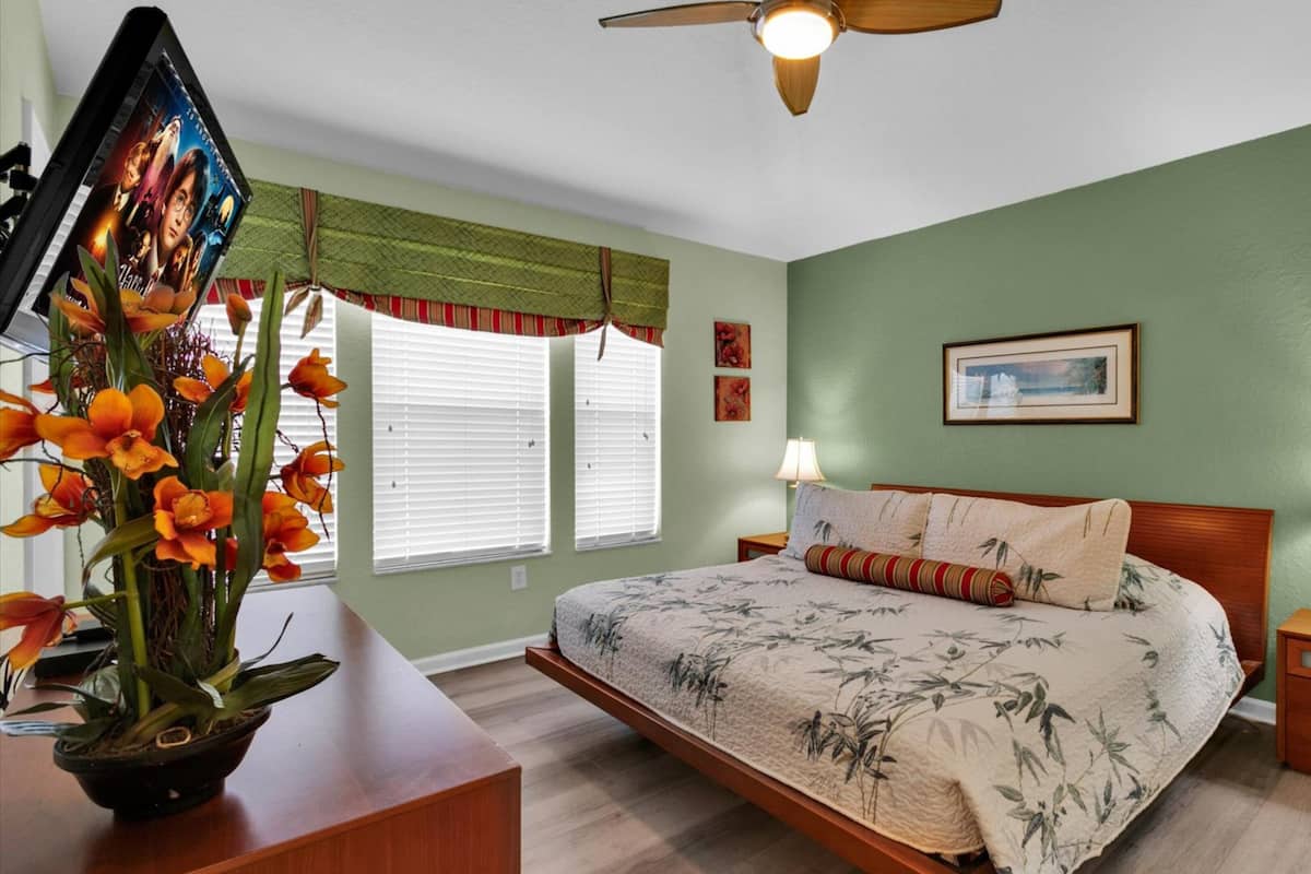 A bedroom with a bed, nightstands, a lamp, a ceiling fan, a window with blinds, and a framed picture on the wall.