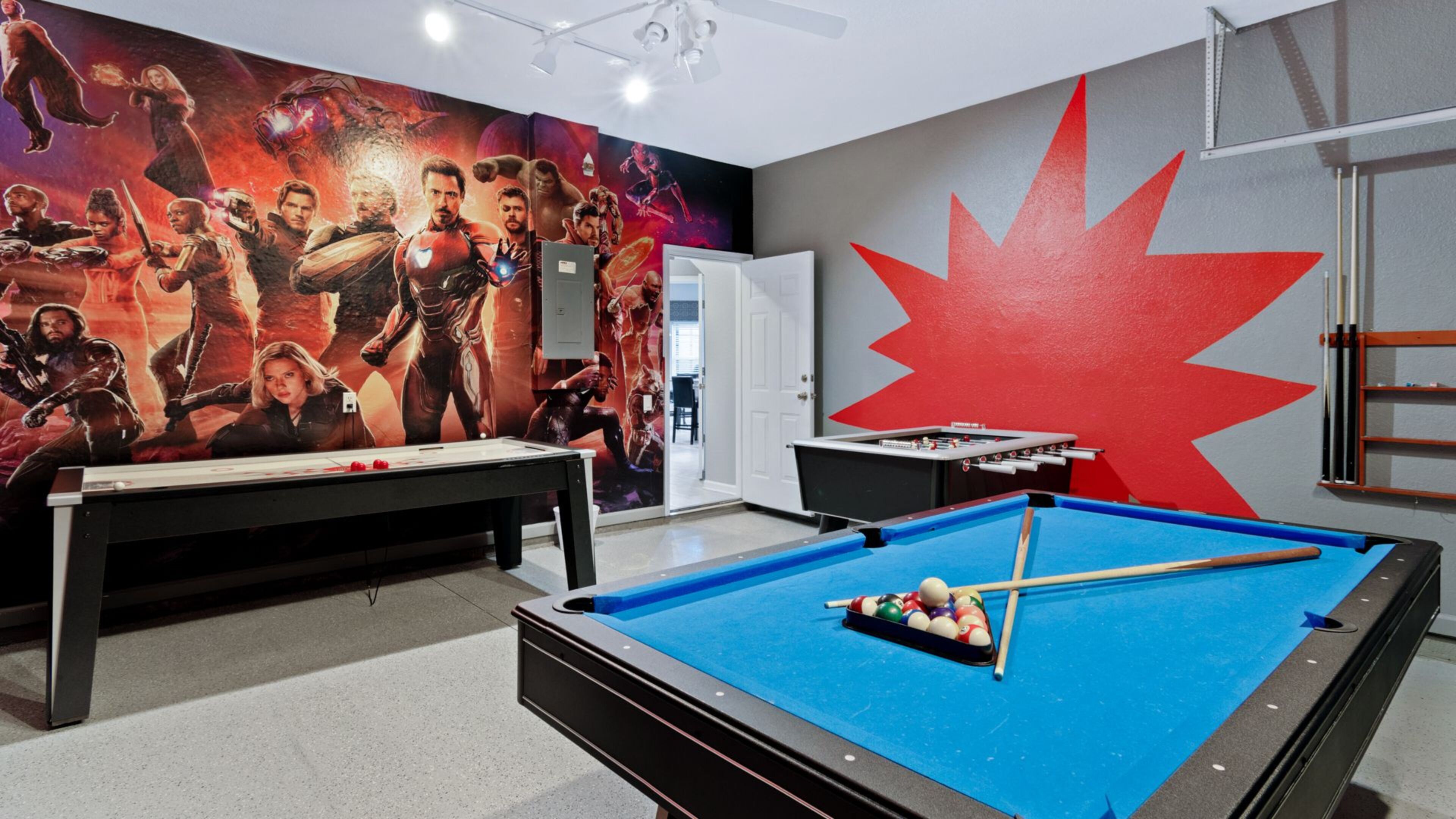 A pool table with a blue felt surface, a foosball table, and a large mural of superheroes on the wall.