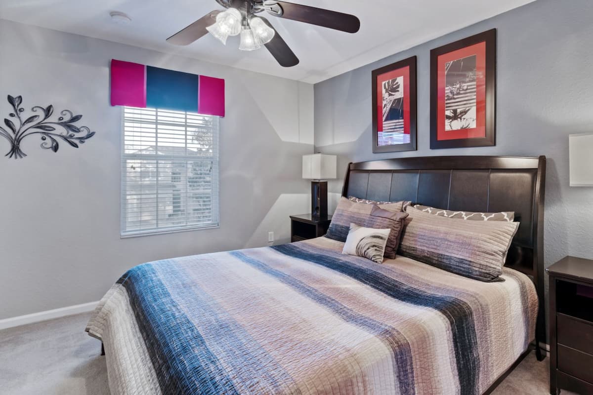 A bedroom with a bed, a ceiling fan, two bedside lamps, a nightstand, a window with blinds, and two framed pictures on the wall.