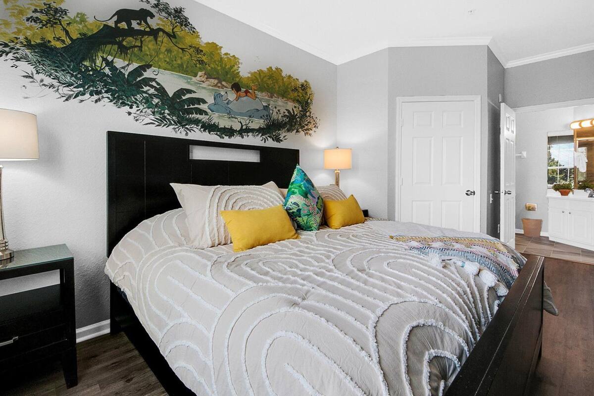 A bedroom with a bed, nightstands, a headboard, a wall mural, and a bathroom visible through an open door.