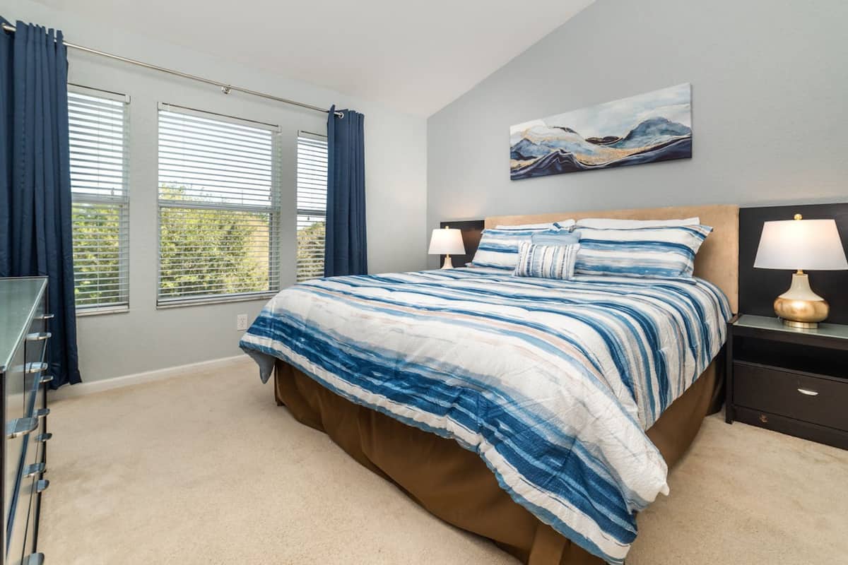 A bedroom with a bed, two bedside tables with lamps, a painting on the wall, and a window with curtains.