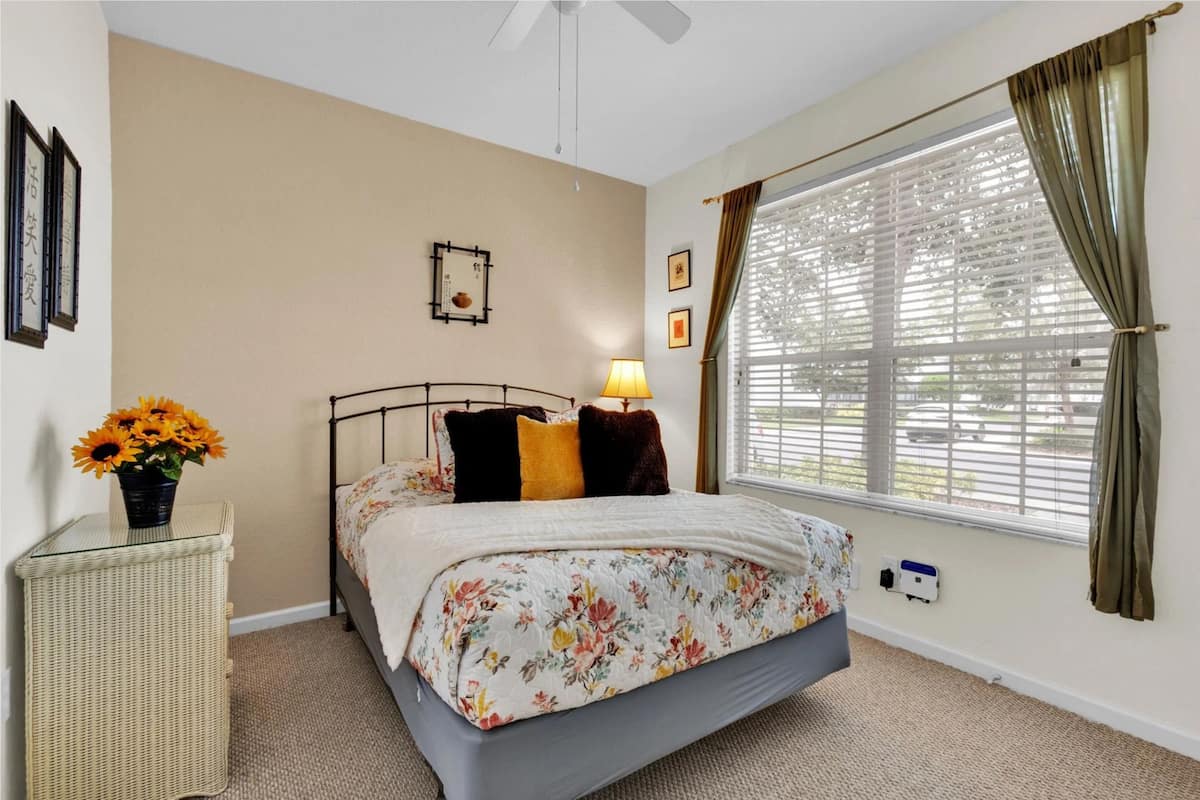 A bedroom with a bed, a bedside table with a vase of flowers, a window with curtains, and a wall-mounted picture frame.