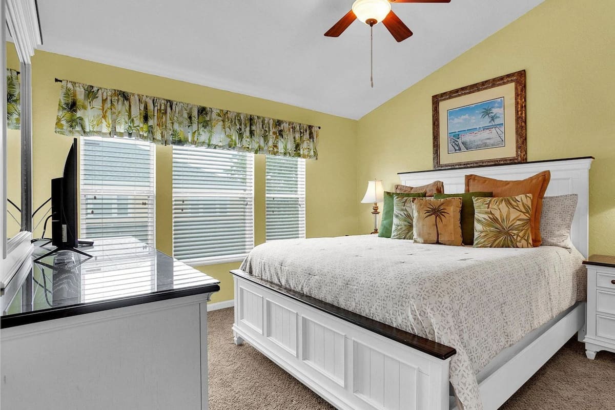 A bedroom with a bed, nightstands, a TV, a ceiling fan, and a window with blinds.