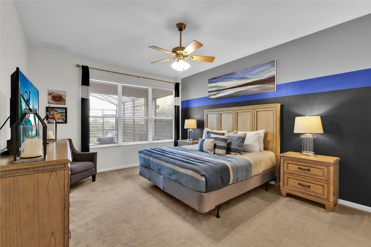 A bedroom with a bed, nightstands, a ceiling fan, a window with blinds, and artwork on the wall.