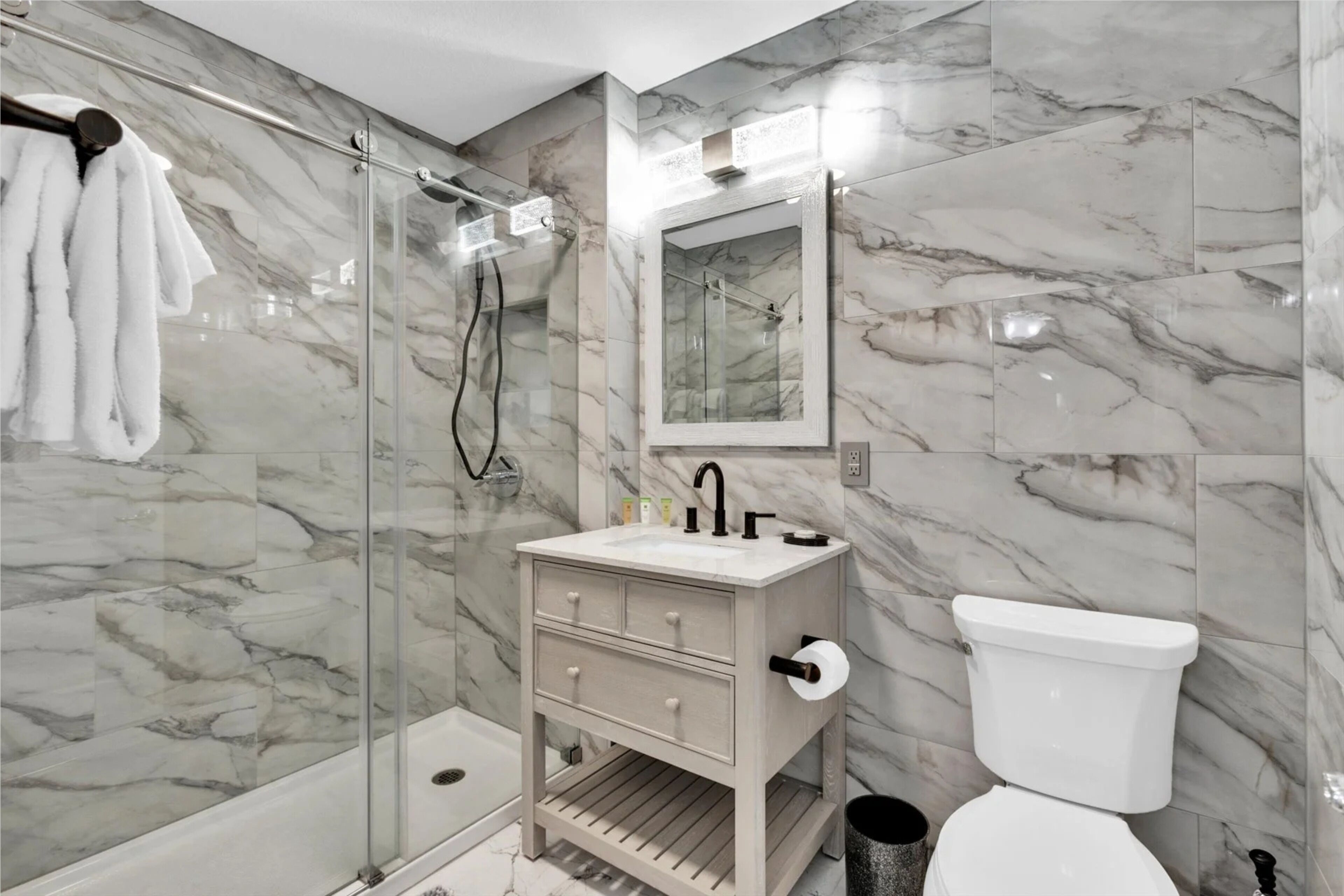 A bathroom with a glass shower enclosure, a marble-tiled wall, a sink with a mirror, and a toilet.