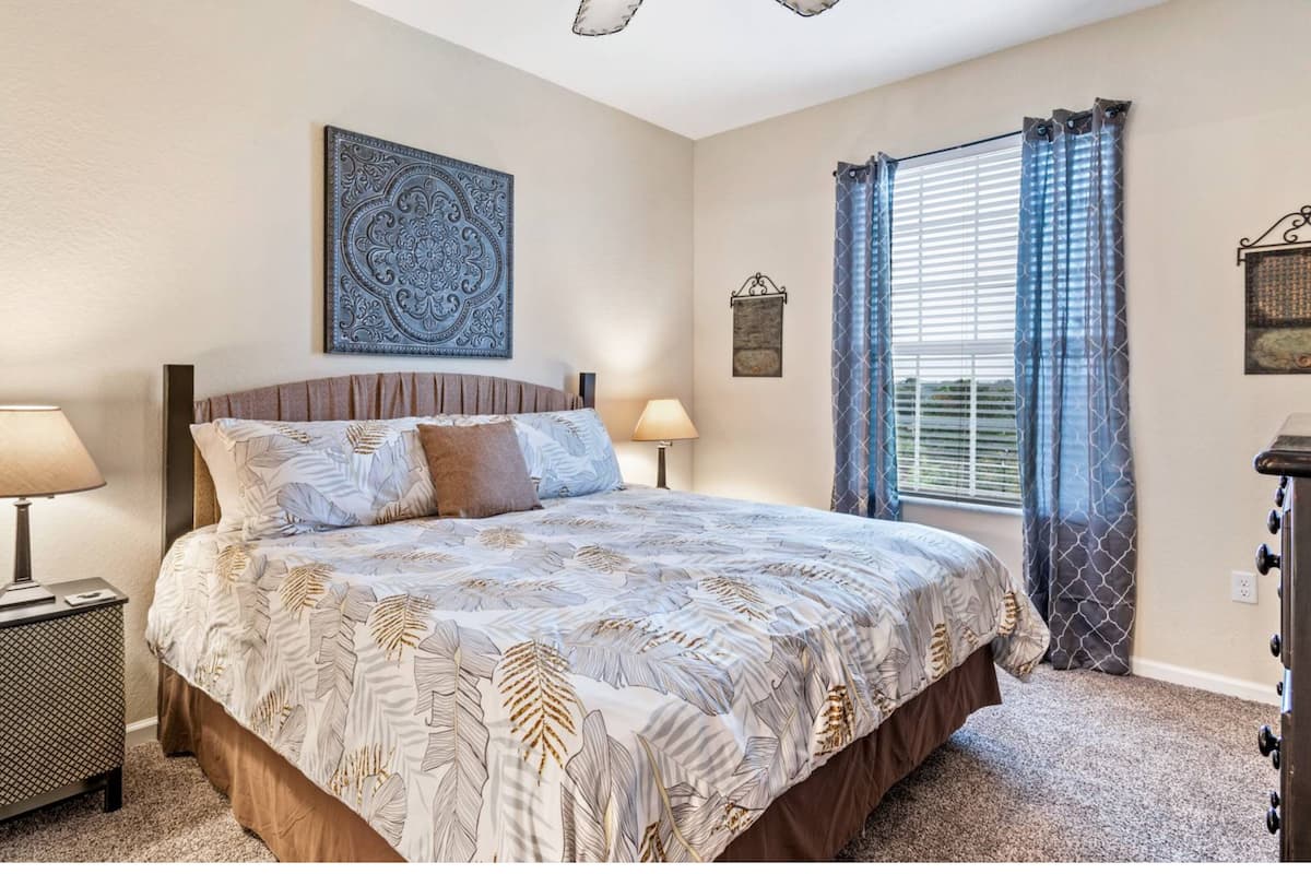 A bedroom with a bed, two bedside lamps, a window with curtains, and a wall-mounted artwork.
