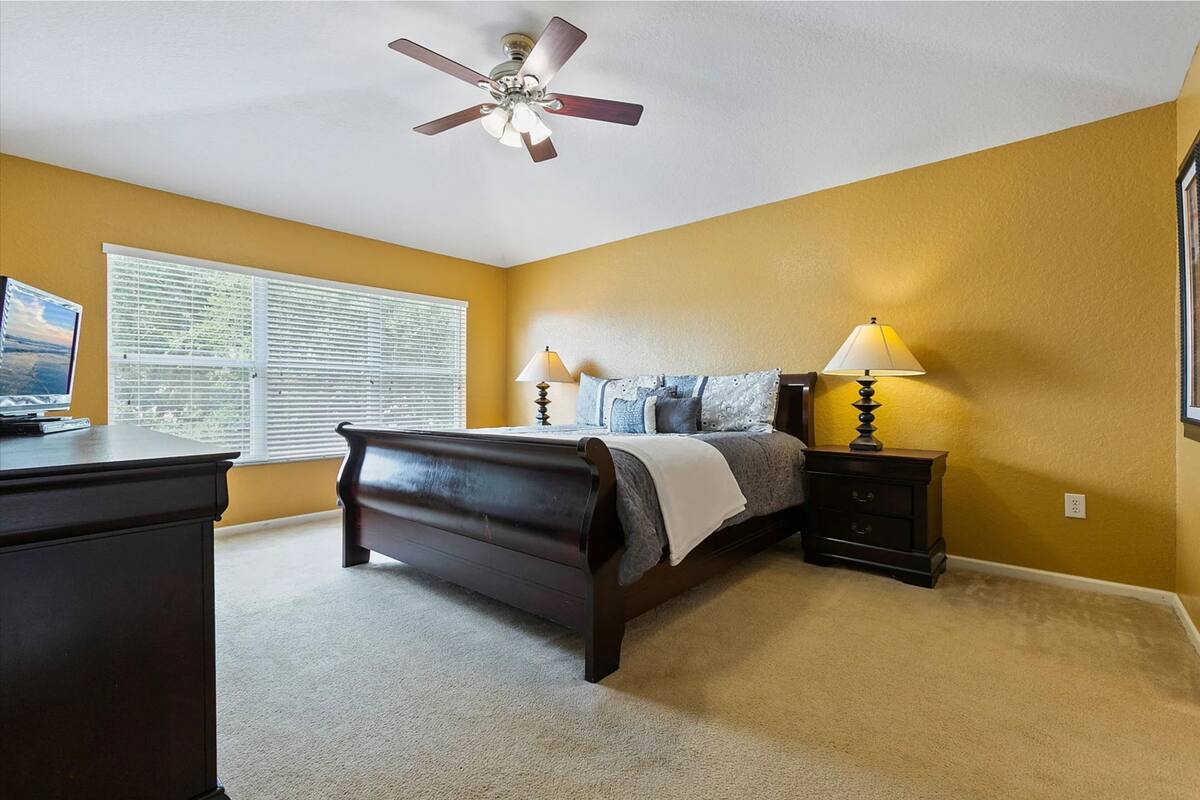 A bedroom with a bed, bedside tables, a desk, a ceiling fan, and a window with blinds.