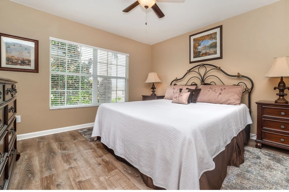 A bedroom with a bed, two bedside tables, a dresser, a ceiling fan, a window with blinds, and two framed pictures on the wall.