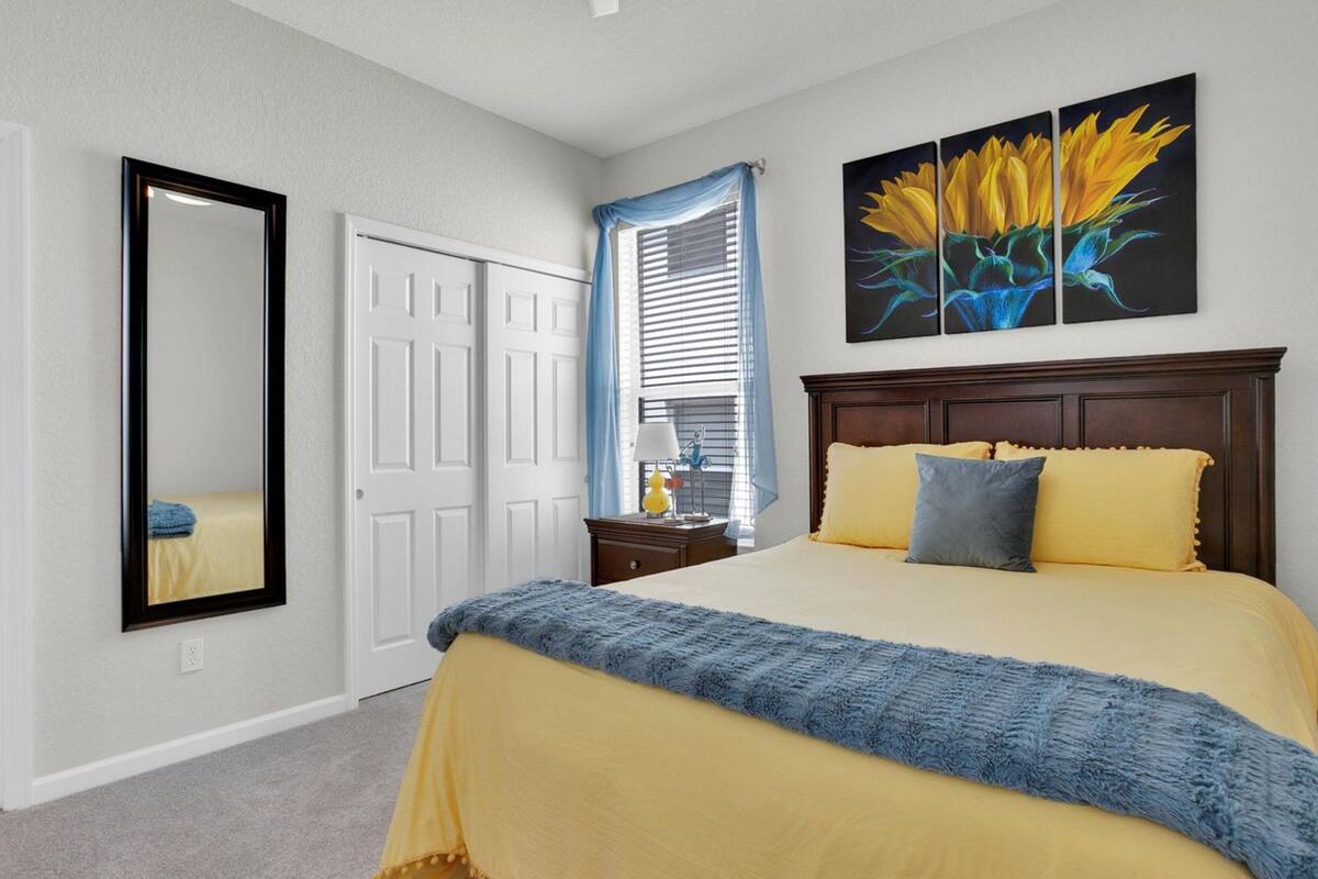 A bedroom with a bed, a mirror, a window with curtains, a nightstand, and a painting.