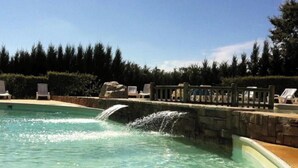 Piscina límpida com cascata, rodeada por espreguiçadeiras e um muro de pedra.