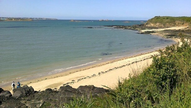 Une plage côtière aux eaux limpides, parsemée de rochers et bordée d’une ligne de côte lointaine.