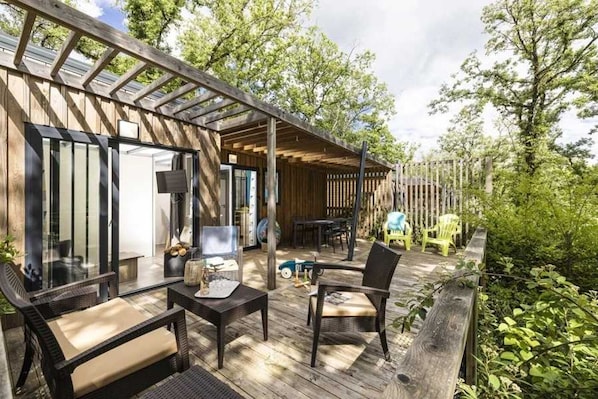 Une terrasse extérieure couverte, dotée d’une table et de chaises, d’une plateforme en bois et offrant une vue sur les arbres.