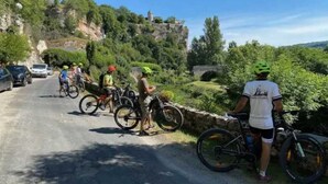 En grupp cyklister på en asfalterad väg med en stenbro och grönska i bakgrunden.