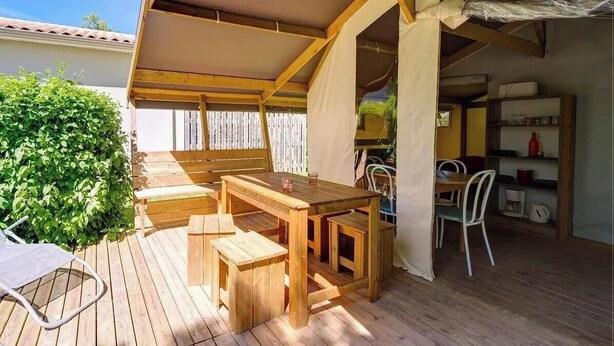 Um deck de madeira com mesa e bancos, uma área coberta com uma cadeira, e um espaço de cozinha com prateleiras e eletrodomésticos.