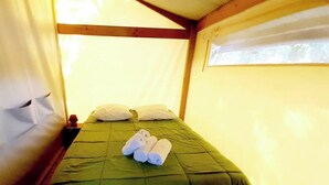 Uma cama de solteiro com roupa de cama verde, duas almofadas e toalhas dobradas em um quarto tipo tenda com vigas de madeira e uma janela.