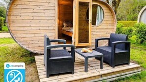 A wooden pod-shaped structure with an open door, a circular window, and a small deck with two chairs and a table.