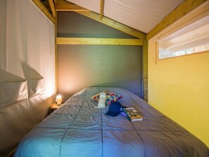 A single bed with a blue blanket, a pillow, a book, and a hat on it. There is a small bedside table with a lamp. The room has wooden beams on the ceiling and a window on the right side. - ZEcamping 3 étoiles familial piscine chauffée pet friendly tente 30m2 (Florac)