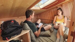Una pequeña cabaña de madera con tragaluz, una cama con almohadas y una mochila en un estante.